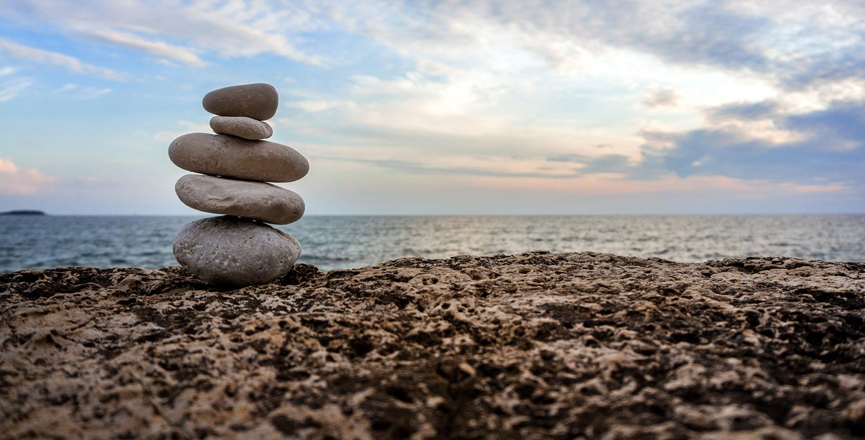 The image shows a rock stack with a tranquil beach scene in the background, featuring a stone wall, ocean, and sky at sunset.
