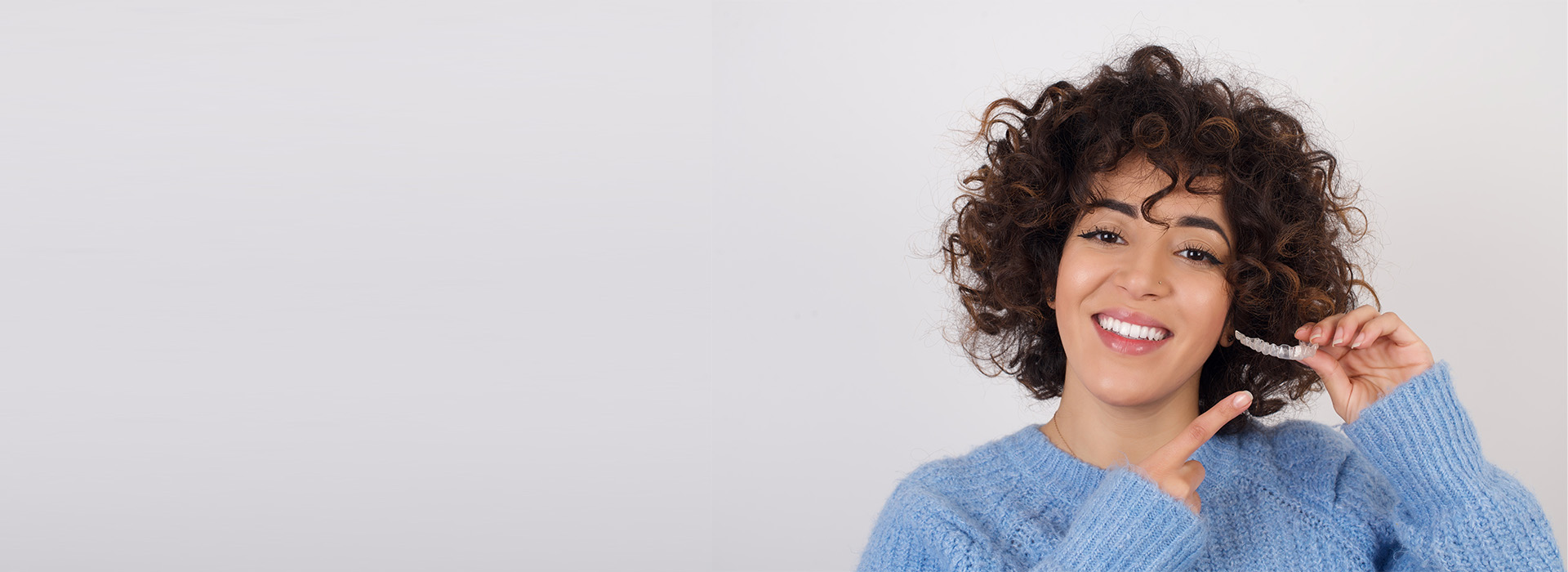 A smiling woman with curly hair brushing her teeth, wearing a blue sweater, against a white background.