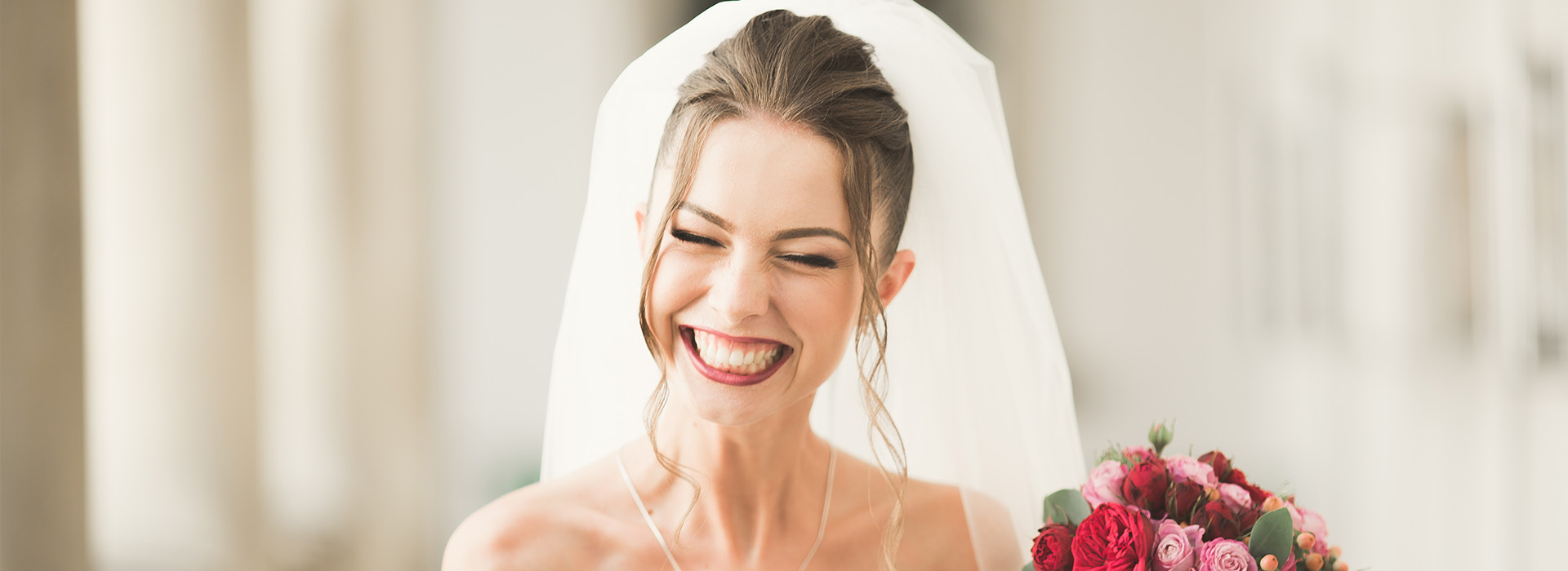 The image shows a woman wearing a wedding veil and smiling, standing next to a bouquet of flowers, with an indoor setting featuring columns in the background.