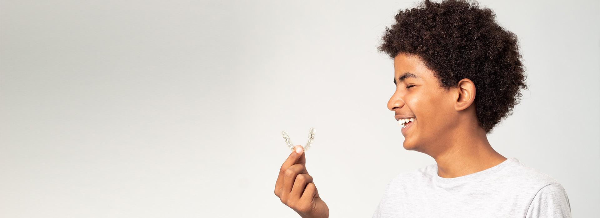The image shows a person with curly hair holding a small object, smiling at the camera against a plain background.