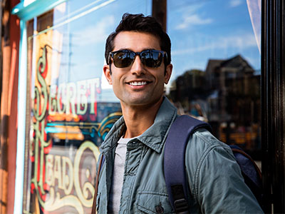The image shows a man standing outside a storefront, wearing sunglasses and a backpack, with a smile on his face.