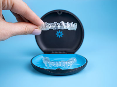 The image shows a hand holding an open box containing a clear tray with a set of dental aligners, which are transparent plastic trays used for teeth straightening.