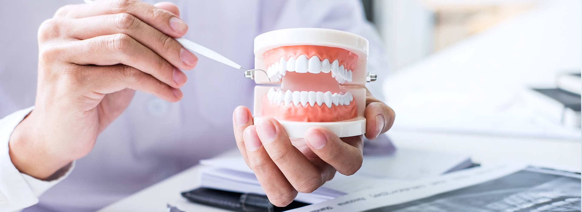The image shows a person holding a toothbrush with toothpaste over a mouth model while another person appears to be inspecting documents, both are in an office setting.