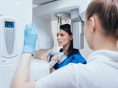 The image shows two individuals in a medical setting  on the left, there s a woman wearing a blue jacket and a headset, standing behind a large, modern-looking piece of imaging equipment, while on the right, another person is partially visible, wearing a white lab coat and a face mask.