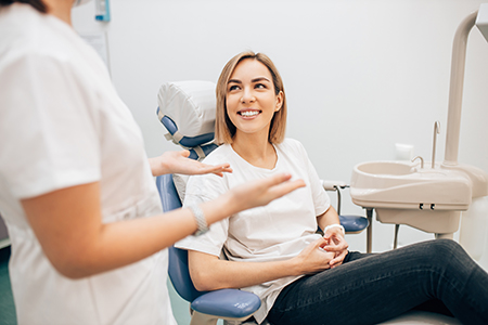 A woman sitting in a dental chair with a smile on her face, receiving dental care from a professional wearing white attire.
