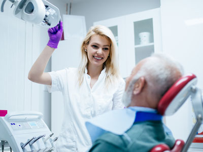 A woman standing next to an elderly man who is sitting in a dental chair, both are wearing face masks, with medical equipment visible in the background.