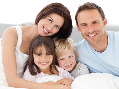 The image depicts a family of five people sitting together on a bed with smiles on their faces, appearing happy and relaxed.